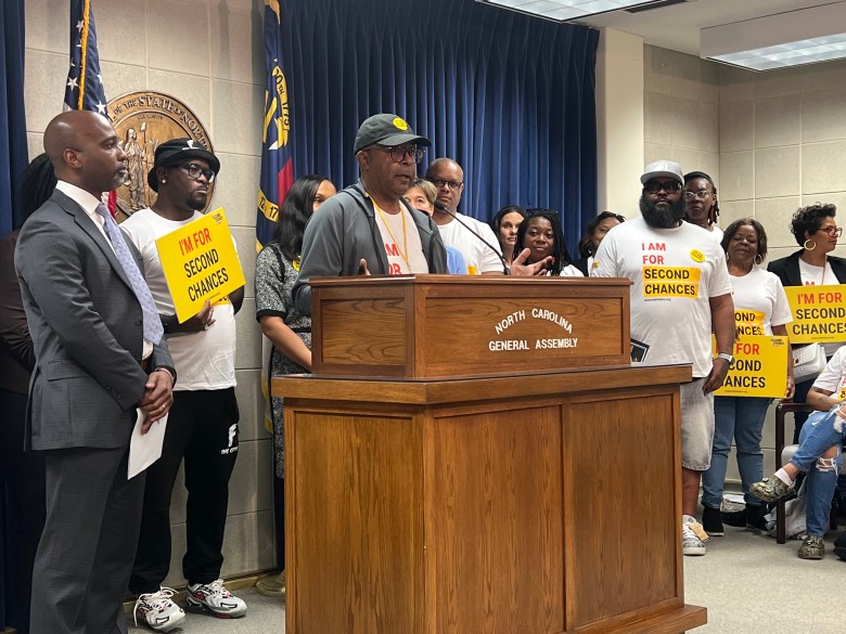 Dennis Gaddy stands in front of a podium with several people standing around him with signs that say 