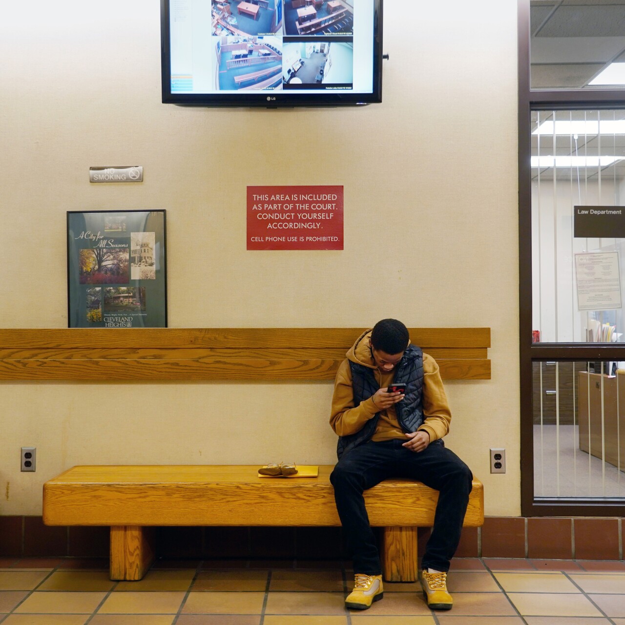 A Black man wearing a dark yellow hoodie and black vest sits on a bench in a court hallway and looks at his phone.