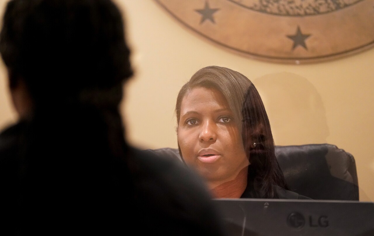 A Black woman with straight hair combed to the side sits at a judge’s bench behind a clear barrier. Another person’s reflection is visible on the barrier.