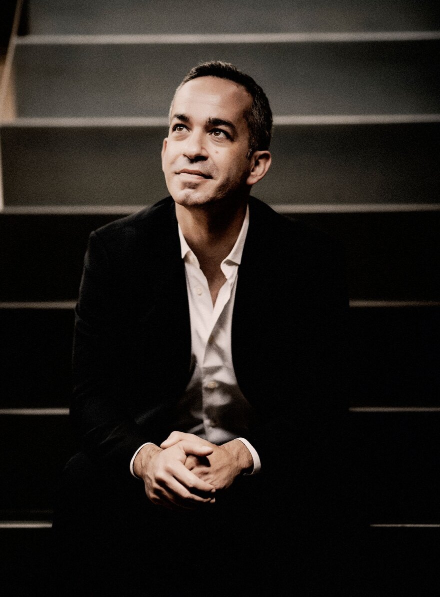 A man sits on darkened stairs, looking up and off to the side with a faint smile. He is wearing a suit, and has his hands loosely clasped.