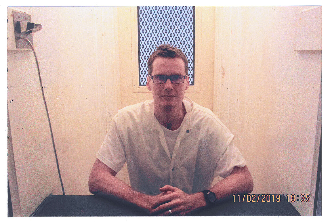 A White man sits at a desk in a small enclosed room with his hands folded in front of him on the desk. He is dressed in a white shirt and he wears dark, thick-rimmed glasses. On the wall to the left, a metal phone cable extends towards the floor. In the bottom right of the photograph, a timestamp label says “11/02/2019 18:35.”