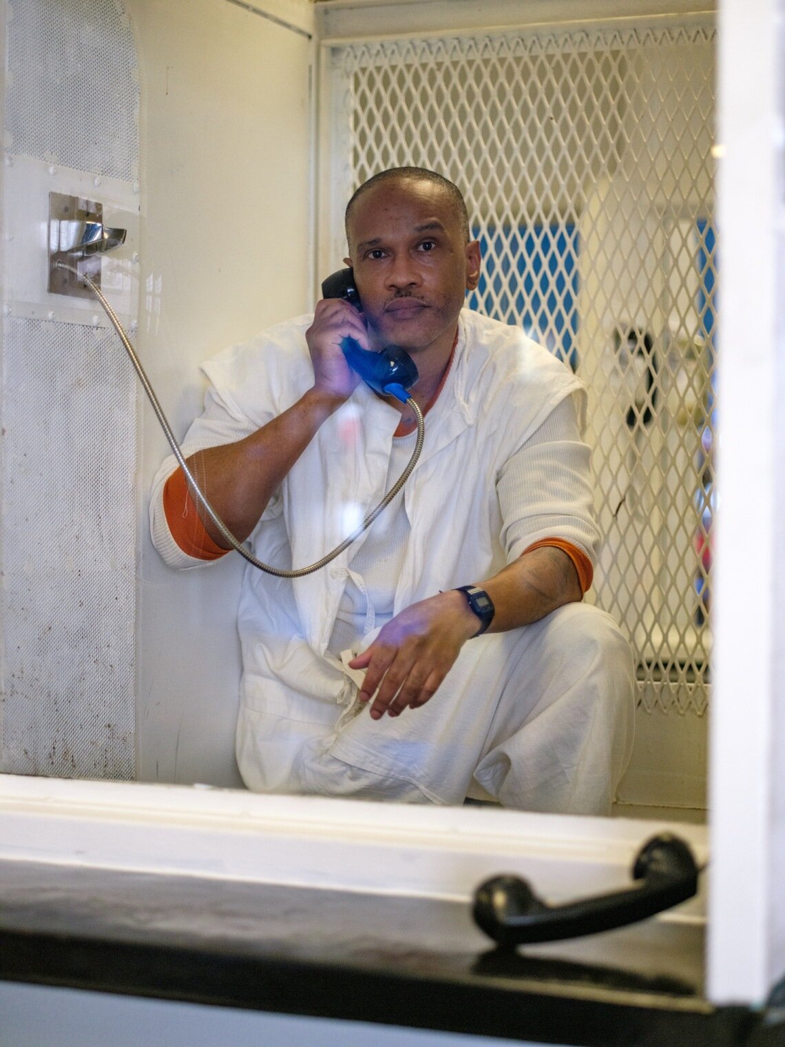 A Black man dressed in white pants and a white shirt stands in a small enclosed room behind a transparent partition with his knee propped up. In one hand he holds a telephone to his ear, with a metal cable connecting the phone to the wall beside him. His other arm is resting on his raised knee. In the bottom right, another black telephone rests on a counter on the foreground side of the transparent partition. 