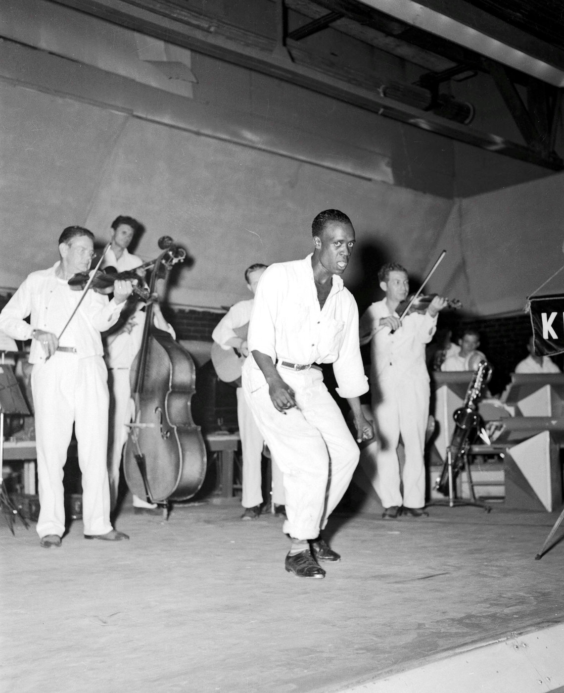 A man faces the camera while dancing in front of a group of musicians playing instruments. 