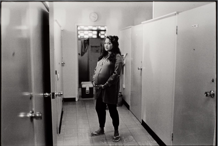 a black and white photo of a young woman showing her pregnancy bump, looking at the camera