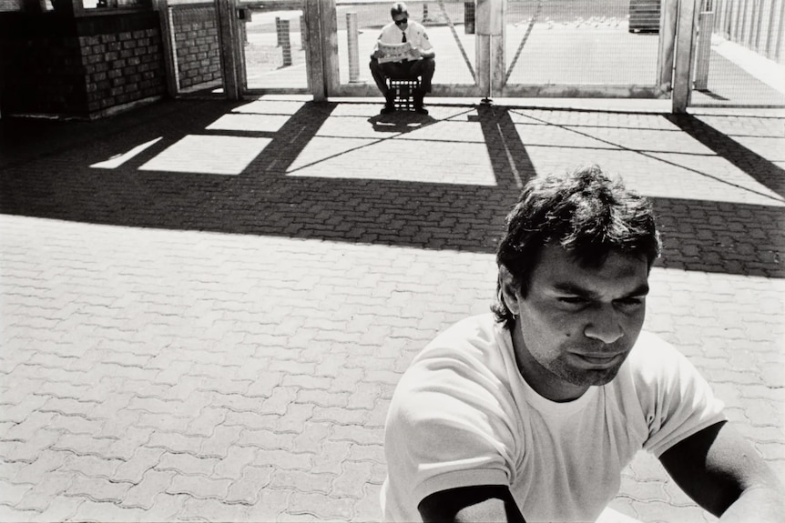 A black and white photo of a male prisoner in the bottom right, as a guard sits on a milk carton and reads the paper