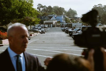 in foreground, man speaks to group. background shows parked cars and small buildings