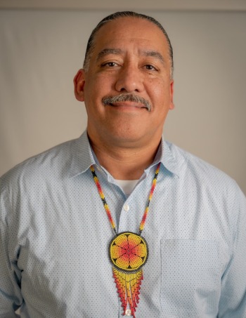 Juvenile justice reform: Headshot of middle-aged Latino man balding with dark hair in light blue shirt with red, orange, yellow and blue beaded medallion necklace, with serious expression, looks into camera.