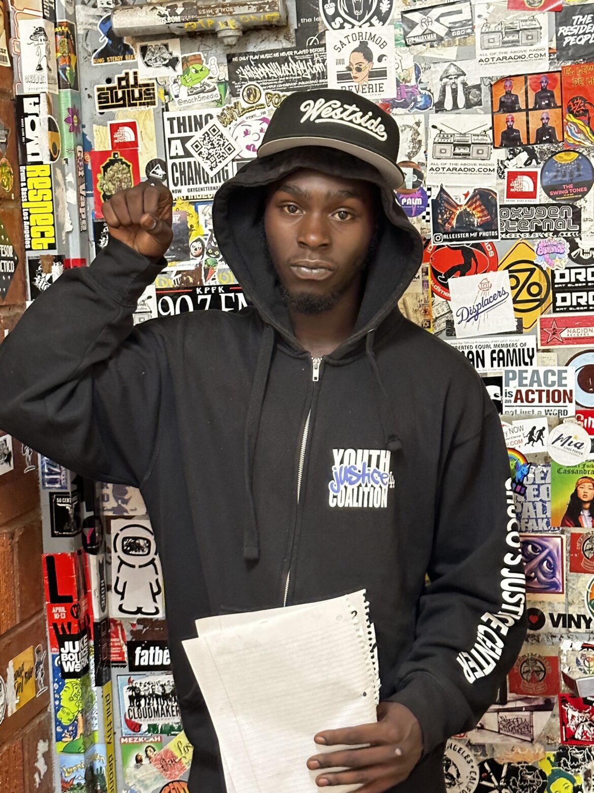 Juvenile justice reform: Black young man in black hoodie holding white papers in left hand leans up against walls completely covered in multi-colored stickers.