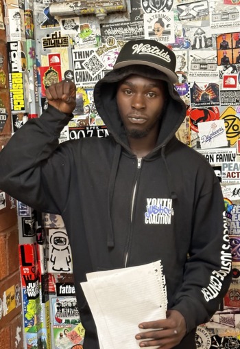 Juvenile justice reform: Black young man in black hoodie holding white papers in left hand leans up against walls completely covered in multi-colored stickers.