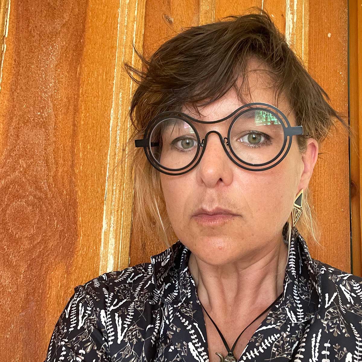 A woman with short hair wearing glasses poses for a photo in front of a wooden wall