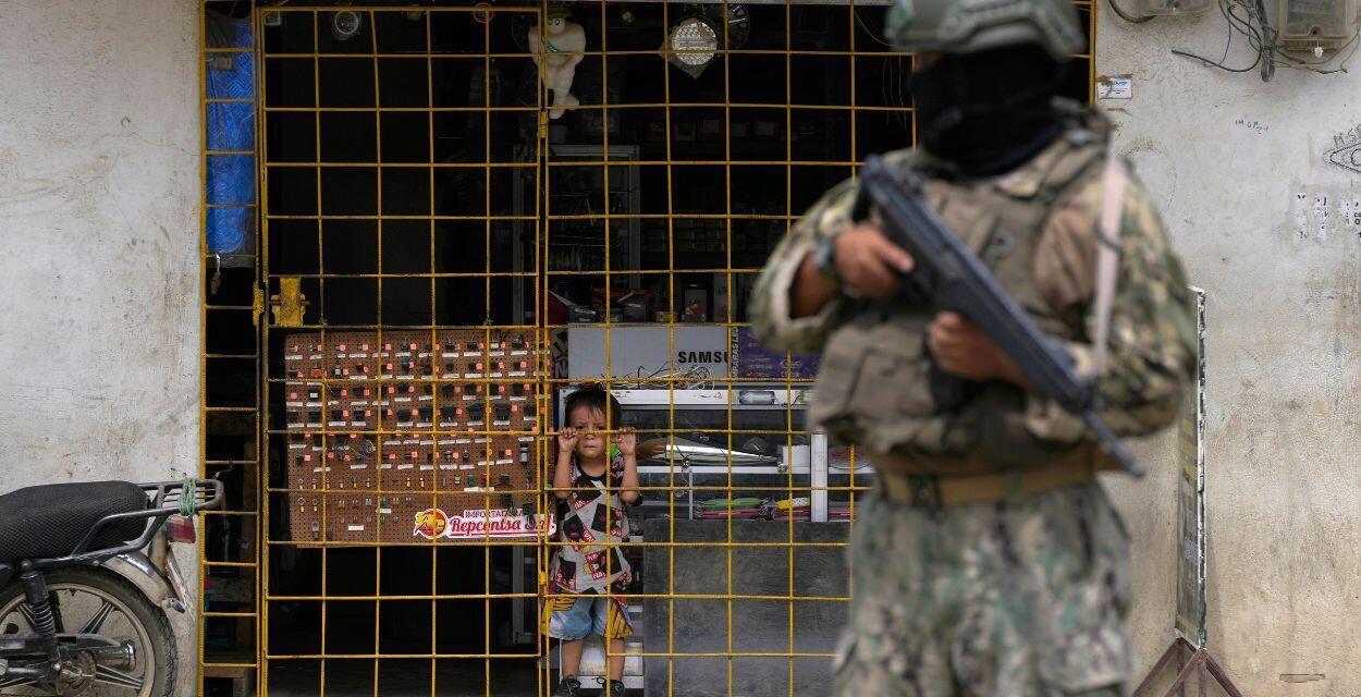 Un niño observa desde detrás de las rejas de una tienda de electrónica abierta mientras un soldado hace guardia en un puesto de control de seguridad colocado por el ejército en Durán, al otro lado de un puente desde Guayaquil, Ecuador, el lunes 14 de agosto de 2023.