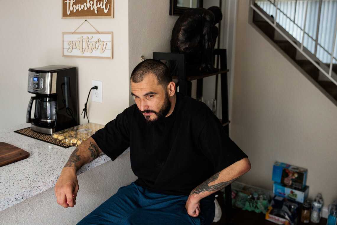 A bearded man with closely cropped hair, a black shirt and tattoos sits at a kitchen island.