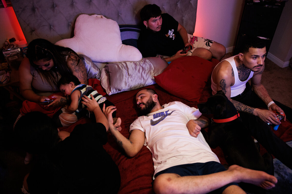 A man wearing a white and blue Nike shirt relaxes on a bed with his relatives. He rests his left hand, which is paralyzed from a past stroke, on his stomach. 