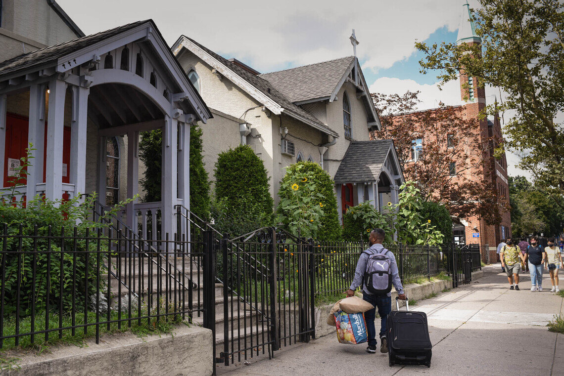 Outside a church on a New York City residential street, a man approaches the front steps, wheeling a suitcase and carrying a shopping bag.