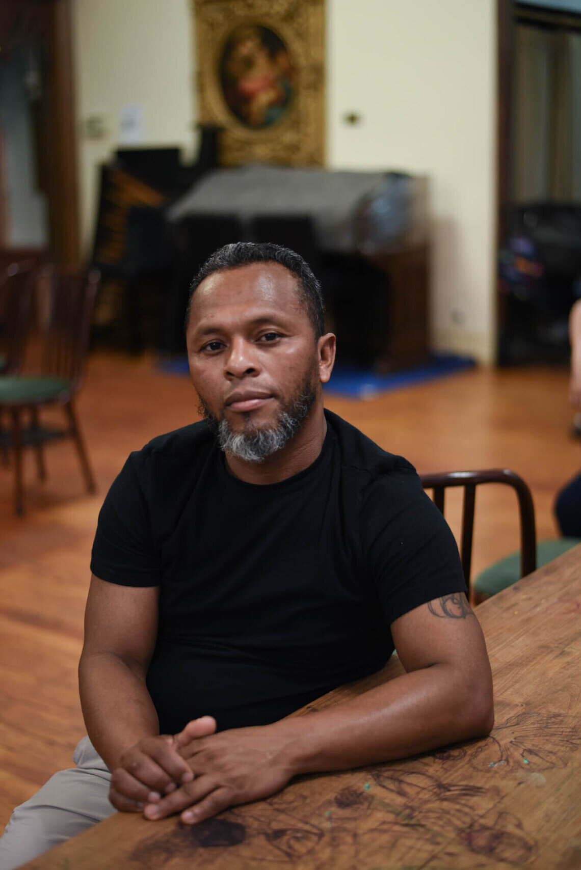 A Venezuelan man with medium-dark skin tone, short hair and a graying goatee sits at a table with his hands folded. He wears a black t-shirt and light gray pants.