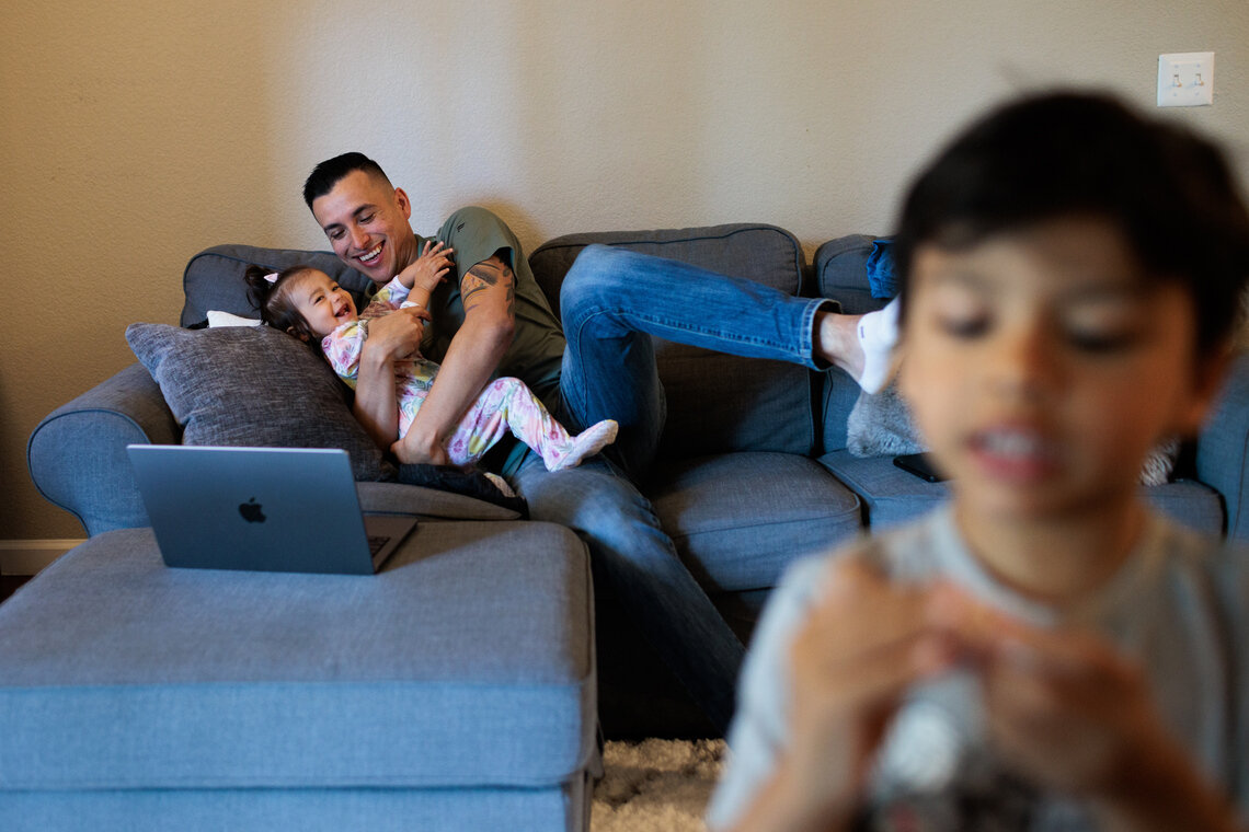 A man wearing a green polo shirt and jeans laughs while hugging his daughter on a blue sectional sofa. 