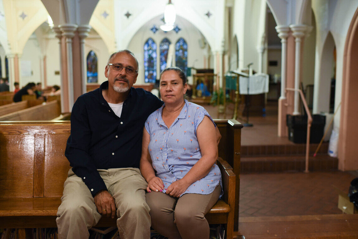 A couple sits next to each other in a church pew. The Colombian man, who is medium-skinned and has a white goatee, wears glasses. He has his arm aroundthe shoulder of a Colombian woman, who is also medium-skinned with dark hair. She has her hair pulled tightly back. They both look forward with neutral expressions. In the background, there are stained-glass windows and other people sitting in pews.