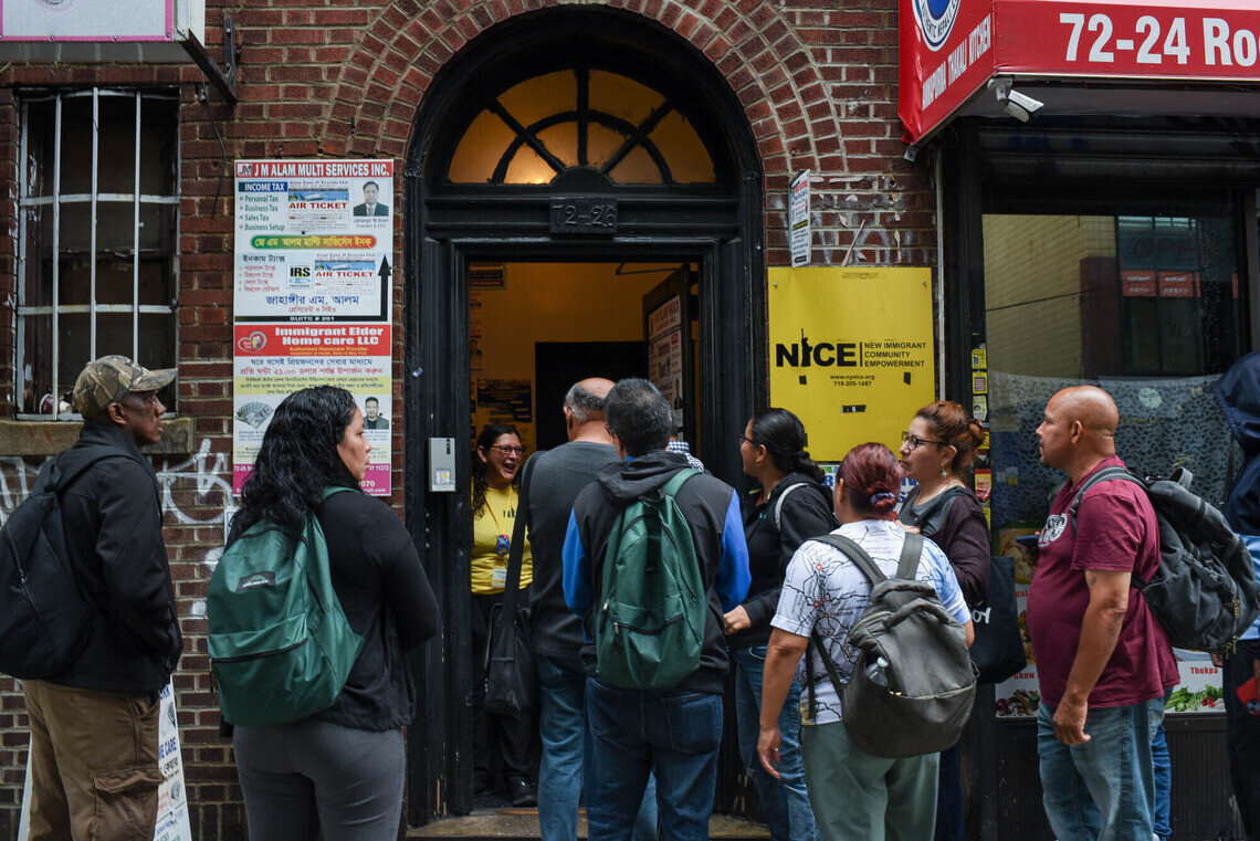 A group of eight people representing various genders and ethnicities wait outside of a building on a street. Many are wearing backpacks. There is a bright yellow sign next to an open door that says “NICE: New Immigrant Community Empowerment.” Inside the doorway, a person in a yellow shirt greets the crowd of people with an open-mouthed smile. 