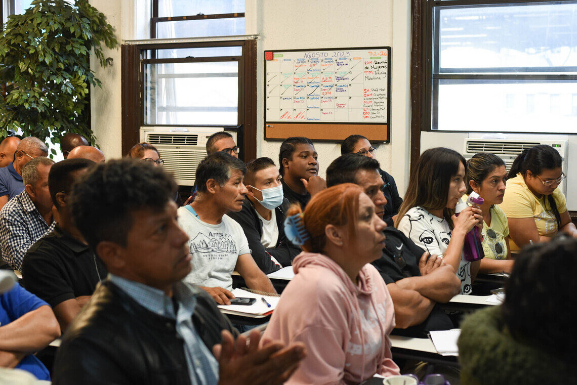 A classroom of more than 15 people representing a mix of genders and ethnicities sits facing in the same direction. Many of the people have focused expressions. In the background, a whiteboard calendar hangs on the wall that says “Agosto 2023.” Many of the dates on the calendar have notations on them.