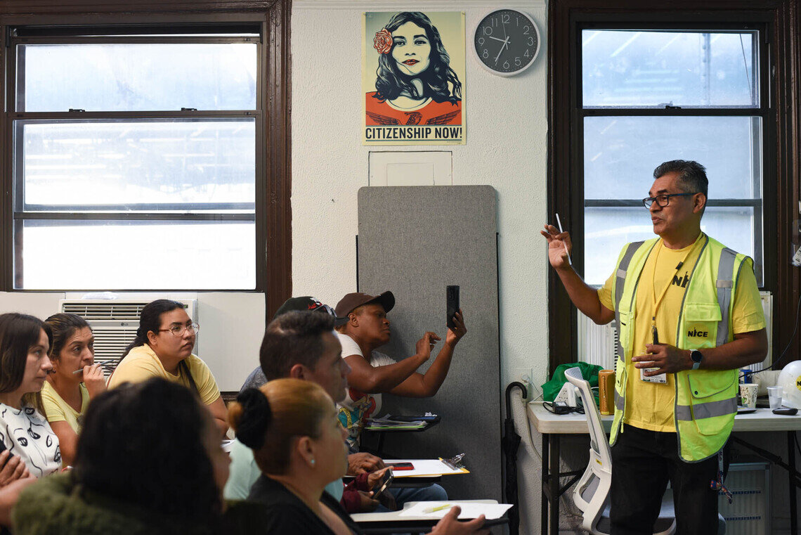 A group of people representing a mix of genders and ethnicities sit in a classroom at school desks. At the front of the room is a man in a yellow shirt wearing a reflective vest that says “NICE.” He has one hand up as he speaks. In the background, on the wall of the classroom, is a poster that reads “Citizenship Now.”