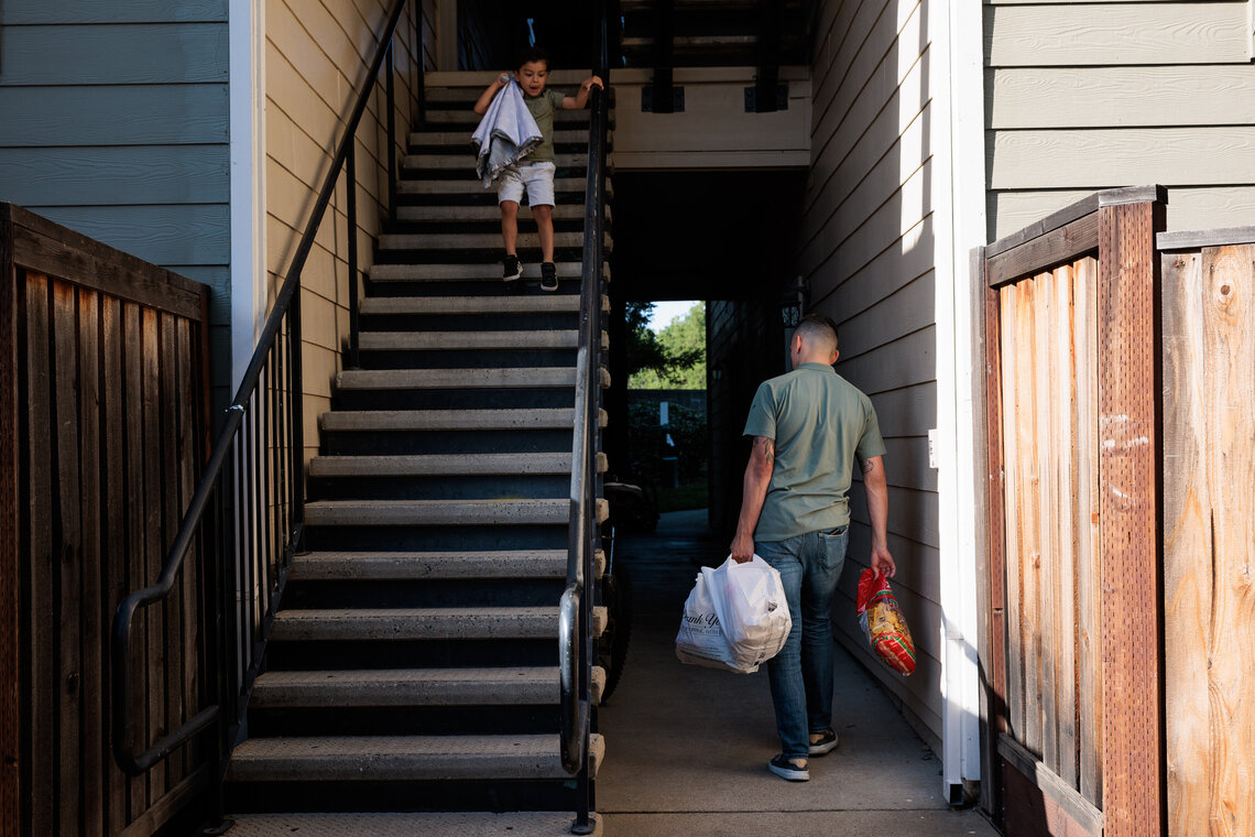 A man facing away from the camera holds two bags of groceries while a child descends an outdoor staircase leading down from the second-floor of an apartment complex. 