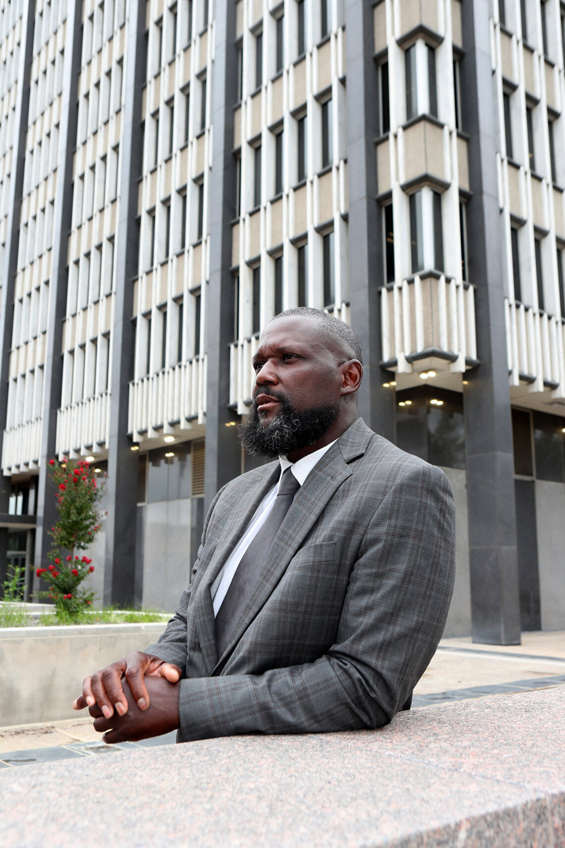 A Black male lawyer, wearing a gray suit, leans his arm on top of a wall for a portrait. A gray and white federal building is in the background. 