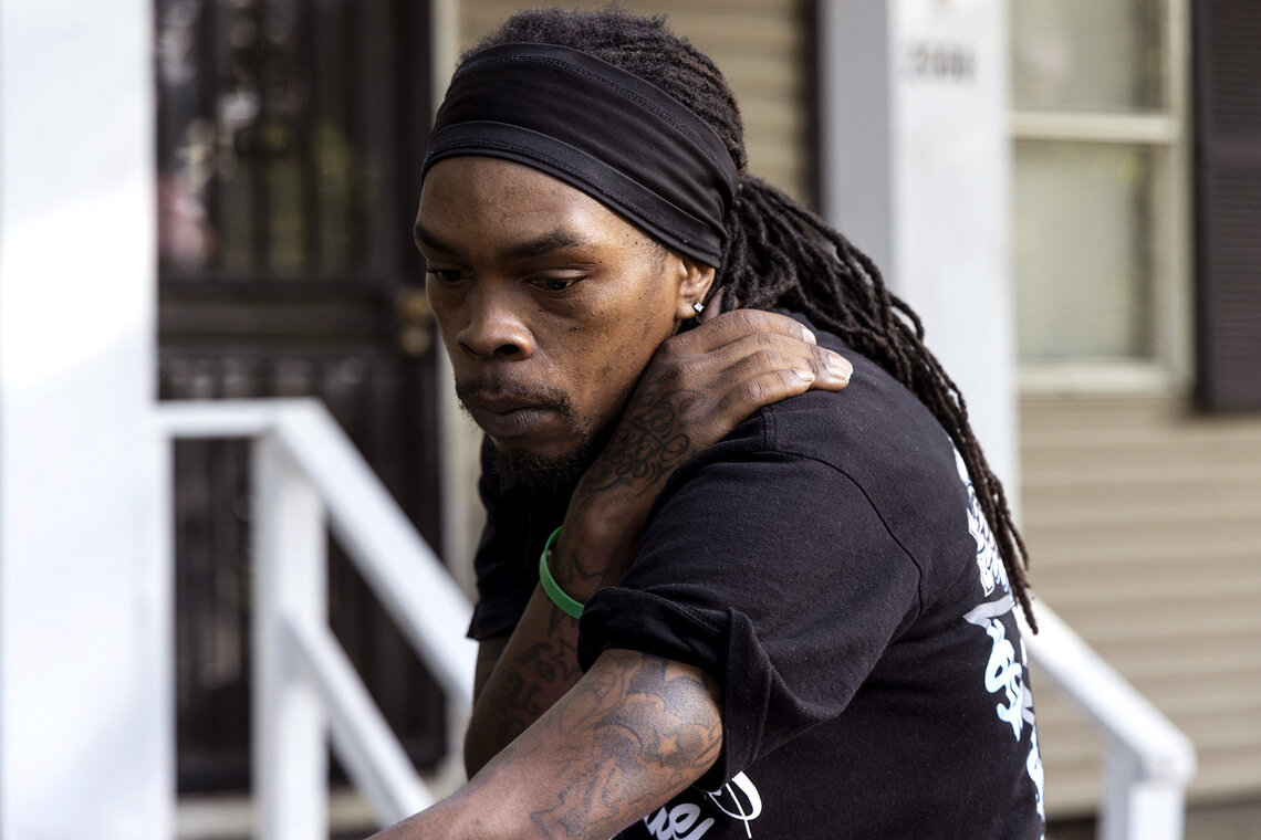 A Black man, wearing a black headband and black graphic T-shirt, leans on his right hand while it is placed on his shoulder. 