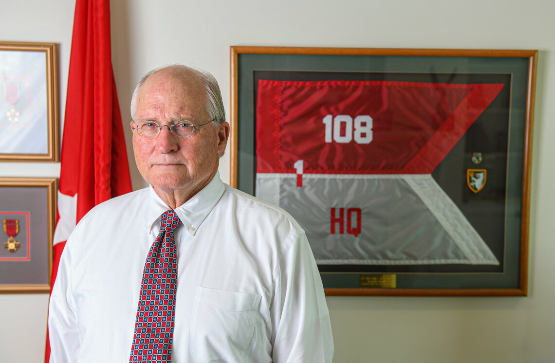 An older White man, wearing glasses, a white button-down shirt, and red tie, standing in front of his framed military memorabilia. 