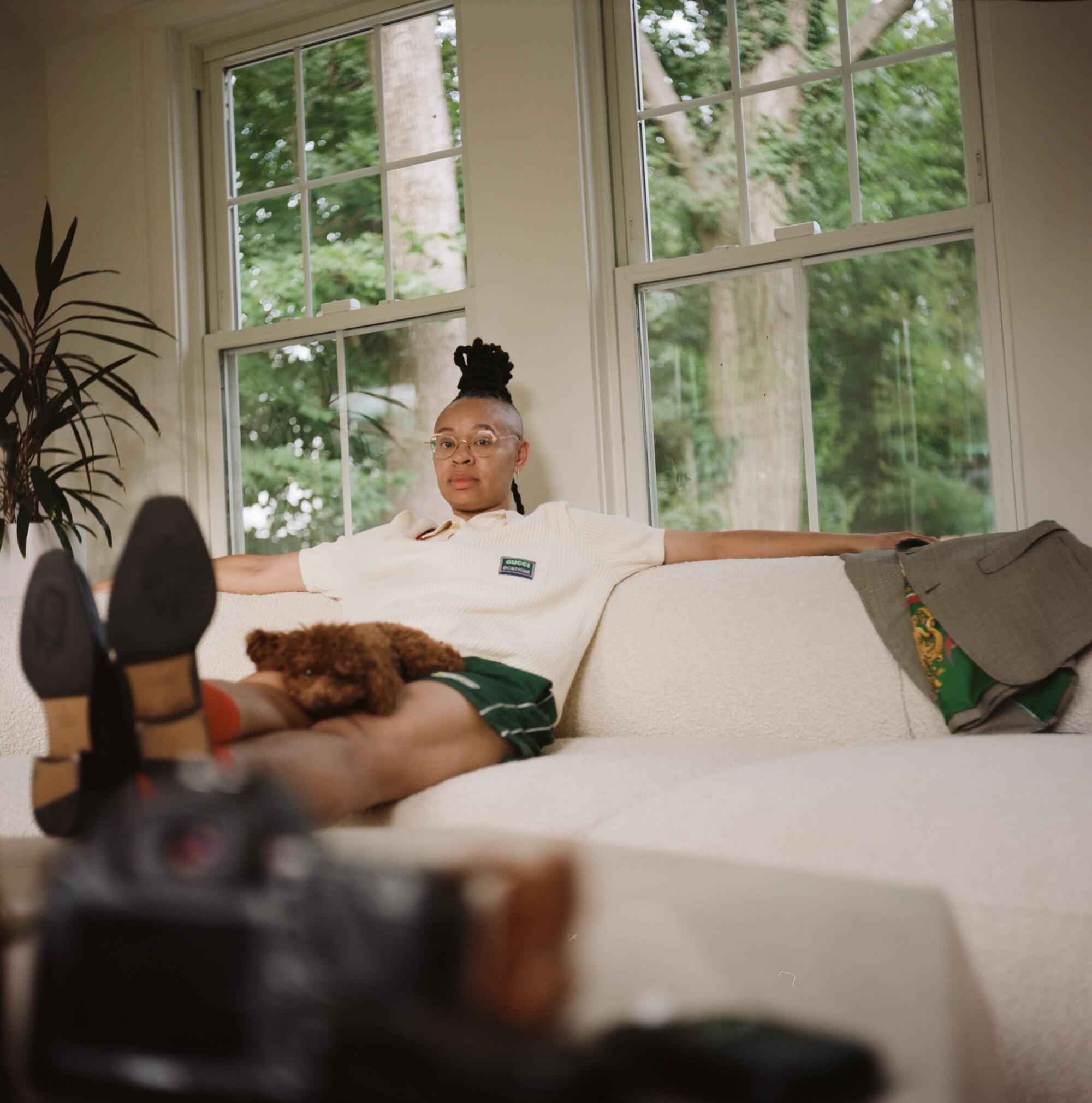 A woman sits on a beige couch, legs propped up on the coffee table, with a dog on her lap. 