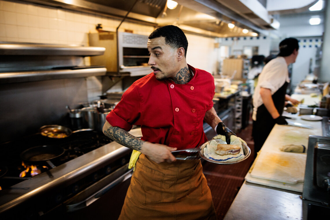 A man with tattoos on his forearm and neck, wearing a red kitchen uniform, transports food on a plate.