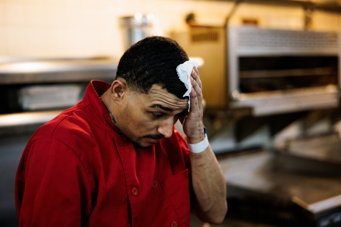 A man with a red kitchen uniform wipes his head with a rag. 