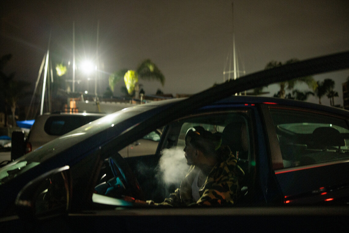 A man smokes in his car at night outside a yacht club. 