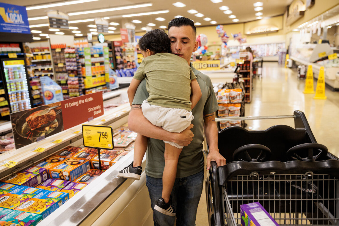 A man wearing a green polo shirt and jeans carries his 4-year-old son while holding a shopping cart in a grocery store. 