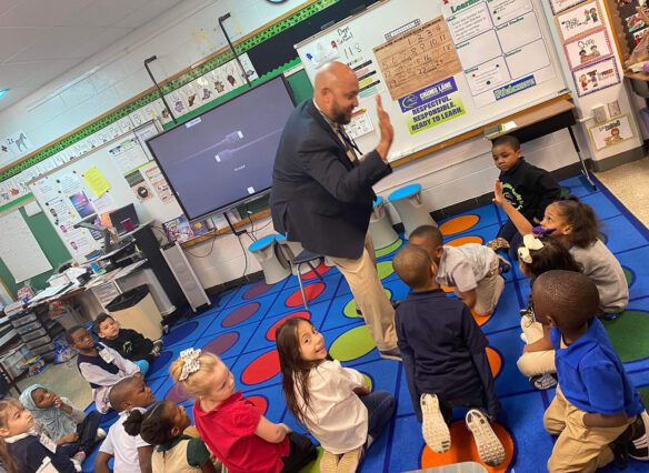 Kumar Rashad high-fives a young student who is sitting with a group of young children on a mat on the floor.