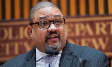 A Black man with a round, closely shaven head, a black-and-white goatee and black-framed rectangular glasses, wearing a dark gray pinstripe suit, white shirt, and light blue tie, looks to the left as he appears to be addressing someone.