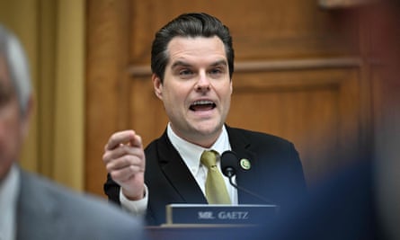 A white man with long brown hair slicked back, wearing a dark suit, white shirt, and dark yellow tie, sits behind a nameplate saying ‘Mr. Gaetz’ as he gestures with his right hand and speaks.