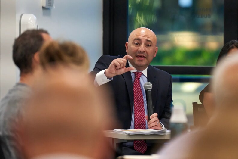 A bald man with light skin tone is seated at a desk, possibly in a classroom, and he's holding a microphone in one hand and gesturing with the other. He's wearing a dark suit jacket, a light-colored shirt and burgundy tie. He's speaking to other people seated in the same room.