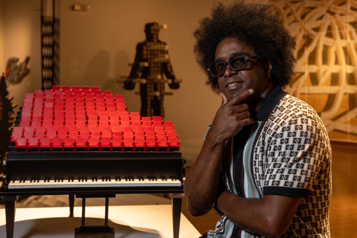 A man wearing a geometric shirt stands in front of  a sculpture in the shape of a piano in a gallery