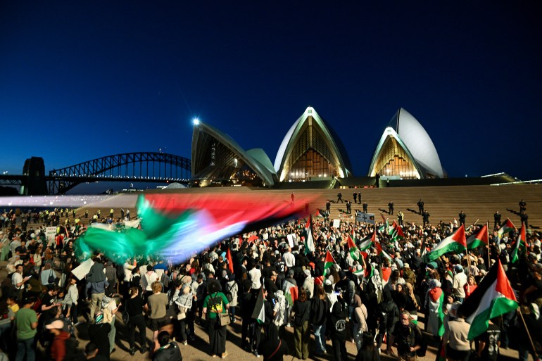 A Palestinian flag waves blurrily in the foreground as people wearing black white red and green are seen in front of the illuminated Sydney opera house against a night sky