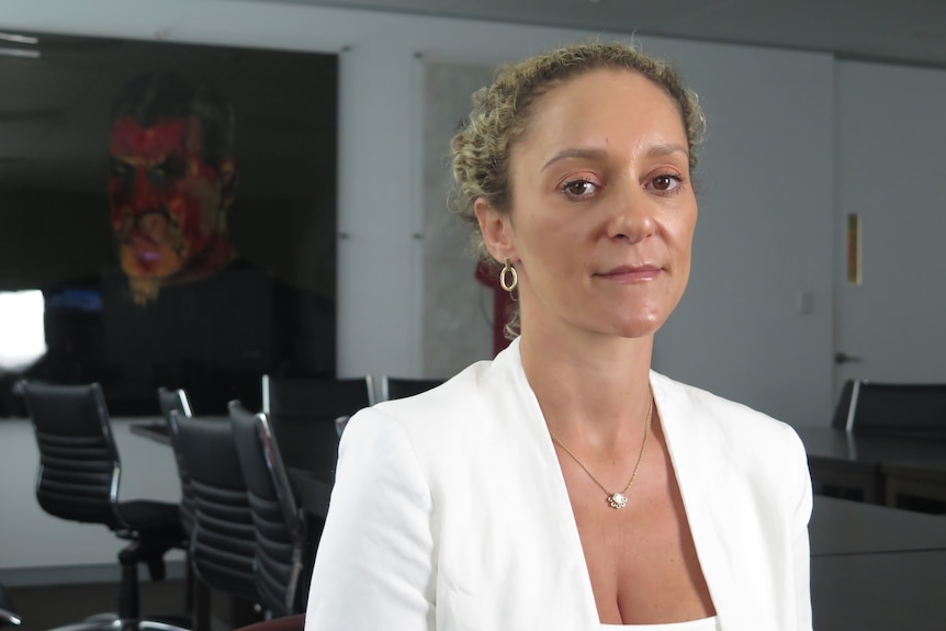 A woman with blonde/brown hair and a white suit, standing in a boardroom