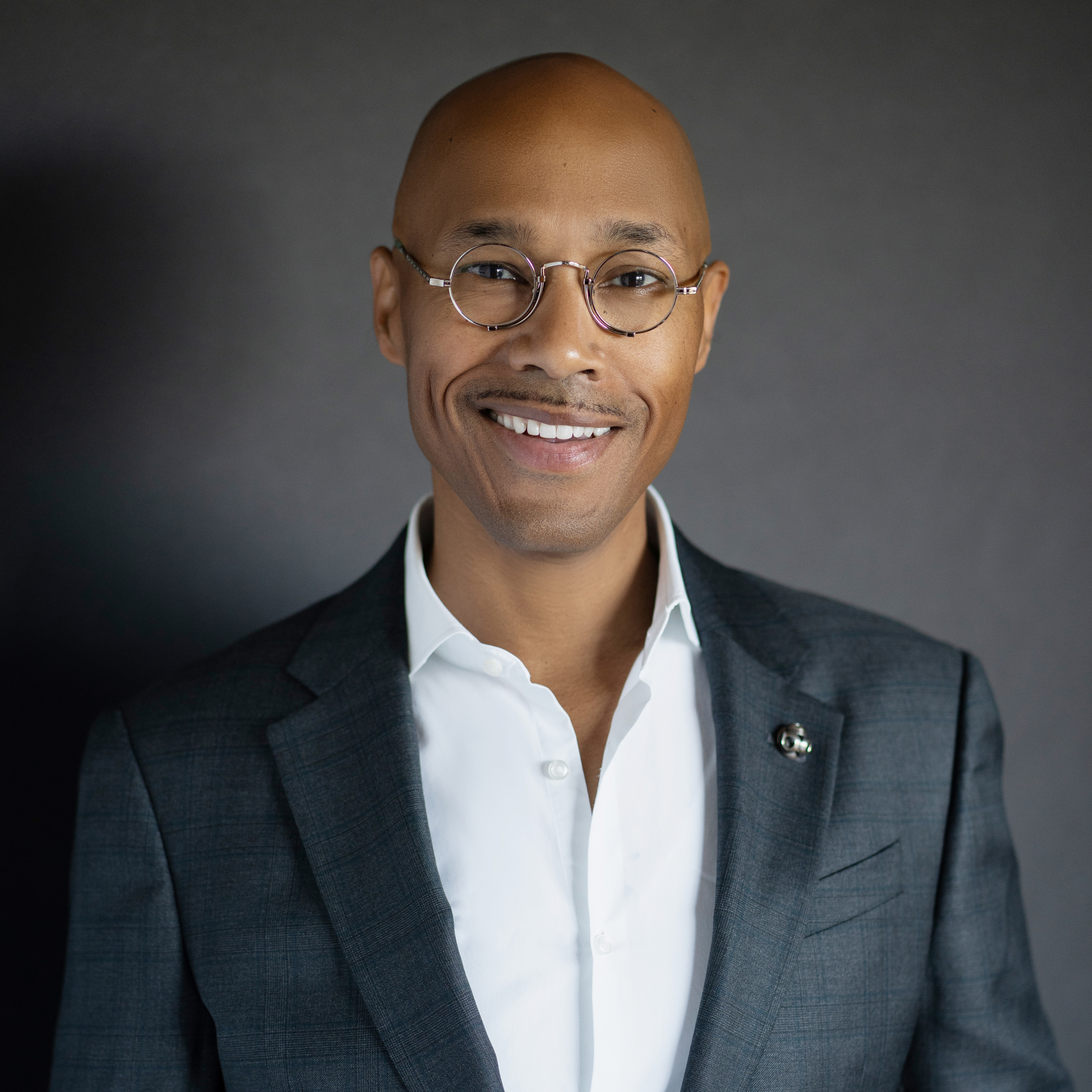 A Black man smiling in formal headshot in white shirt and gray suit, no tie, round glasses