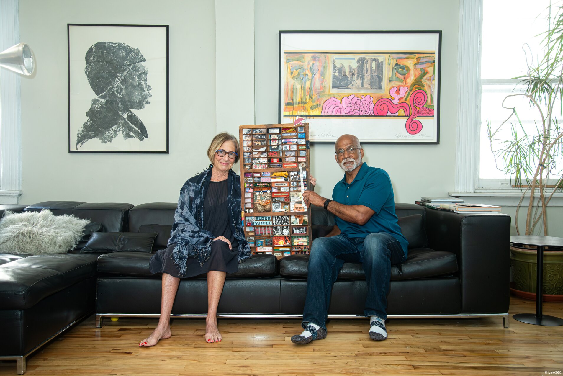 A middle aged white woman and a middle aged Black Hispanic man sit on a sectional couch, holding a piece of artwork. Two other pieces of artwork are on the wall behind them.