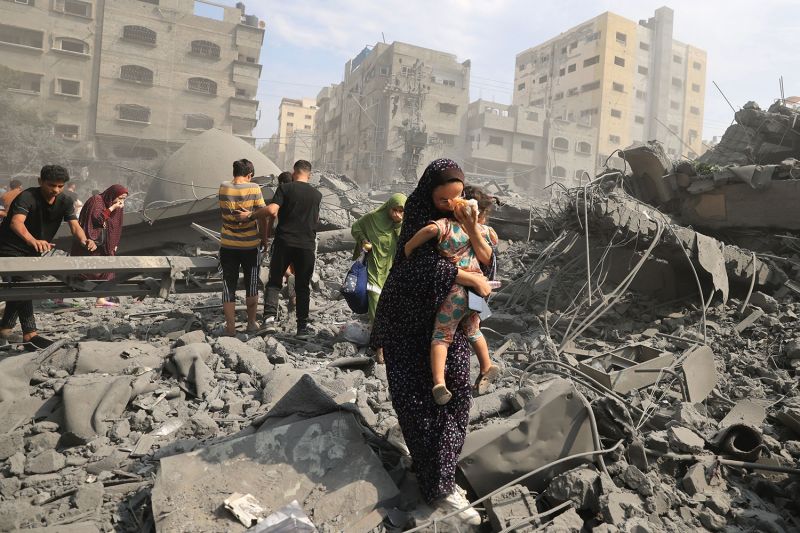 A woman wearing a headscarf and dress carries a small child as she and others walk across rubble following an Israeli airstrike on the Sousi mosque in Gaza City. Dust, smoke, and destroyed buildings fill the landscape.