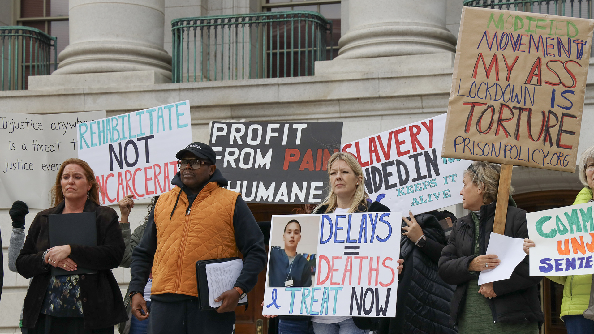 People stand in front of a building with marble masonry and pillars and hold signs that read 