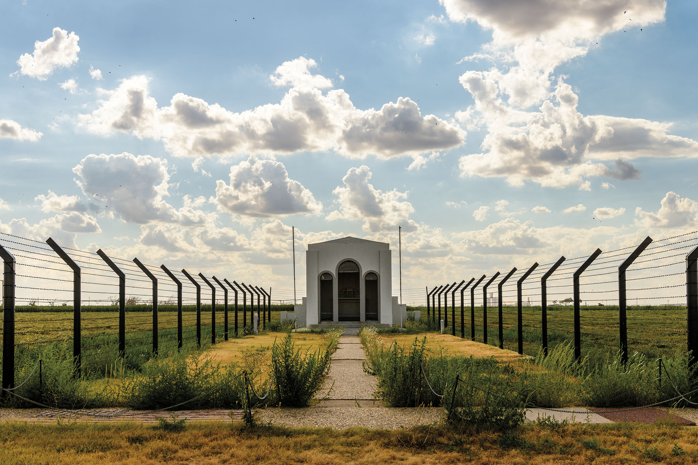 A view of a white chapel flanked by tall wire fences and scrubgrass