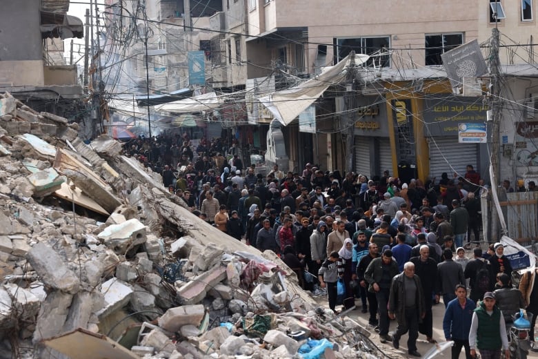 A large crowd of people walk past rubble of a destroyed building.