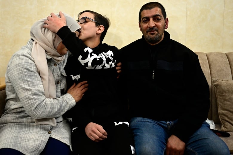 A teenage boy in glasses sits on a couch between a man and a woman. He kisses the woman on the forehead as the man smiles into the camera. 