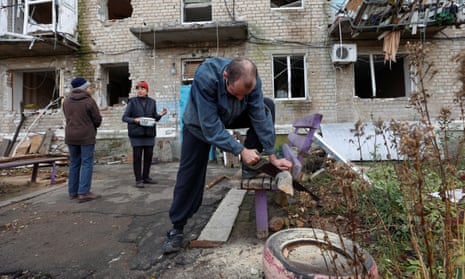 A man saws firewood next to his bombed-out apartment building as two women talk in the background. 