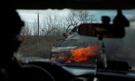 Photo taken from inside a car through the windscreen showing the back of a soldier driving and a burning car in the centre of the frame. 