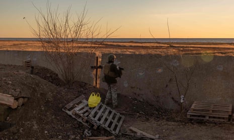 A Ukrainian soldier stands next to a wall at dusk, a cross and yellow plastic carrier bag are in the foreground of the image. 